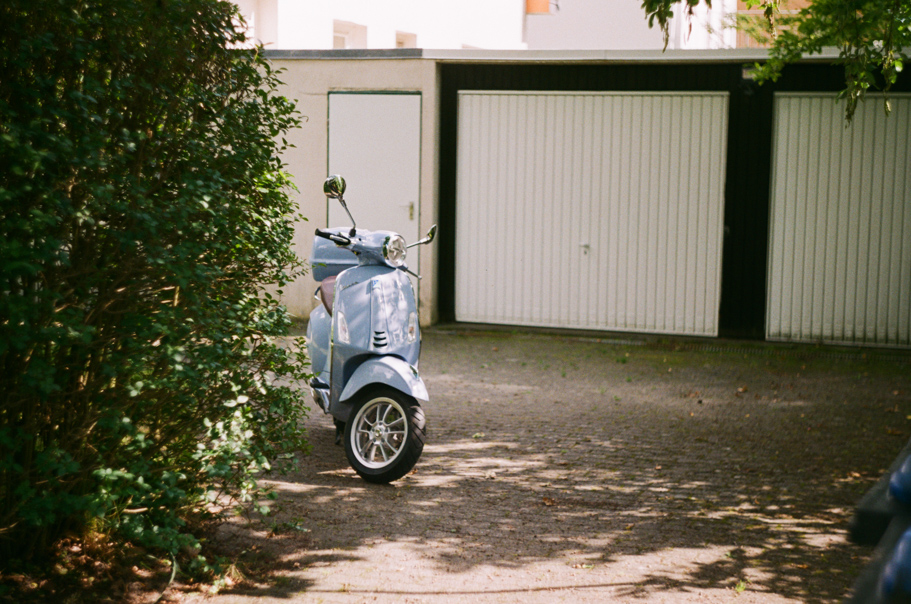 A light blue Vespa standing in a drive way with a green bush on one side and garage door on the other side. Shot on Fujifilm Fujicolor C200.